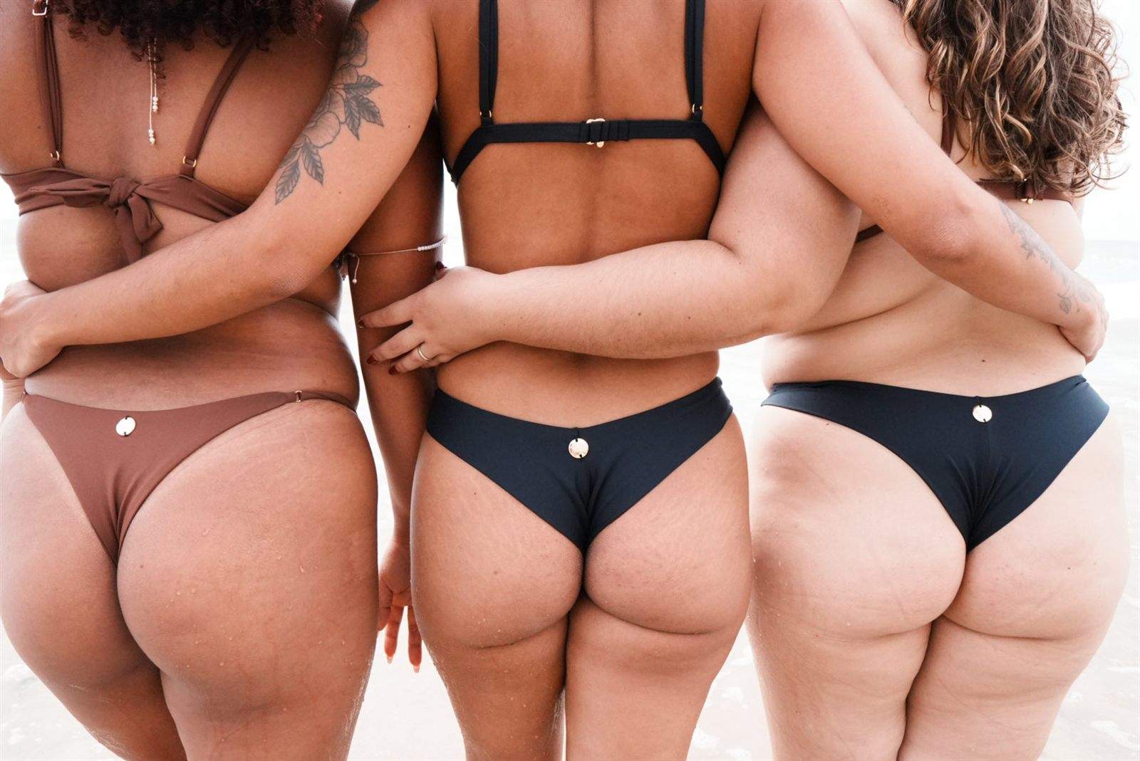 Três mulheres de biquíni, abraçadas de costas para o mar